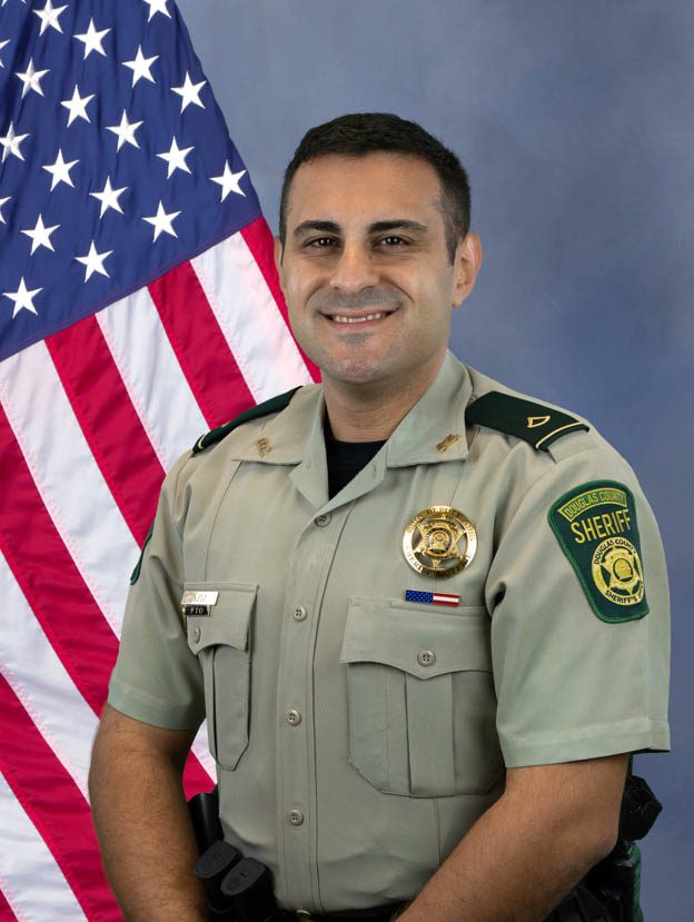 Portrait of a Douglas County Sheriff officer in uniform standing in front of an American flag, smiling.