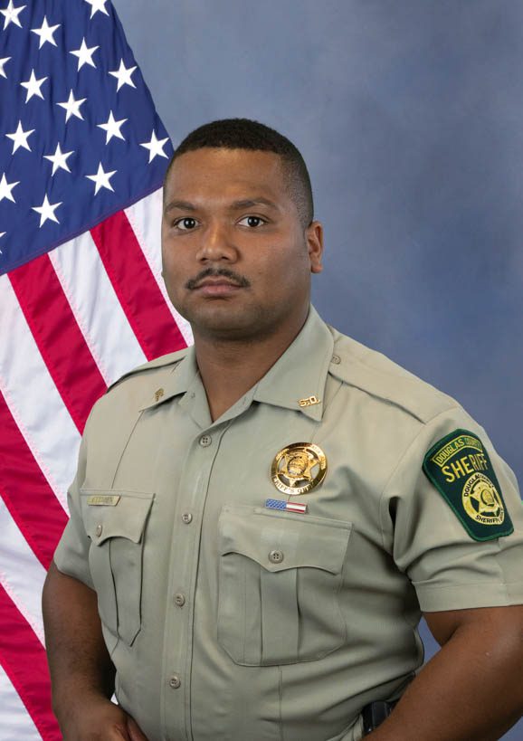 Portrait of a Douglas County Sheriff deputy standing in front of an American flag, wearing a uniform with badges and a shoulder patch labeled 'Douglas County Sheriff'.
