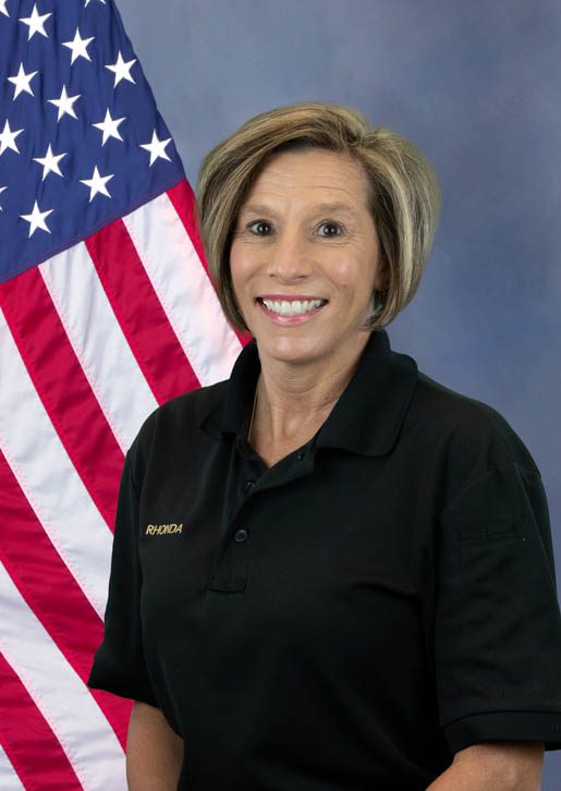 Portrait of a woman smiling in a black shirt with the name 'Rhonda' embroidered, standing in front of an American flag.