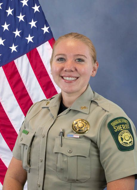Uniformed female sheriff's deputy smiling with American flag in the background, wearing badge and Douglas County Sheriff patch on uniform.