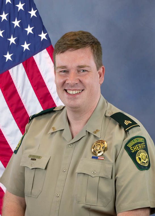 Portrait of a Douglas County Sheriff deputy in uniform standing in front of an American flag, smiling at the camera.