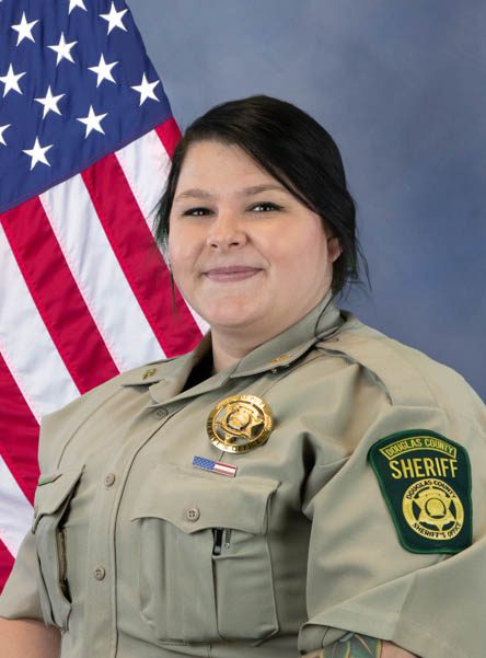 Portrait of a female sheriff's deputy in uniform with a Douglas County Sheriff badge, standing in front of an American flag.