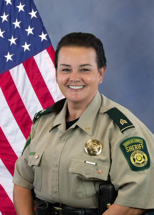 Portrait of a Douglas County Sheriff’s Office deputy sergeant smiling, wearing uniform with badge and patches, standing in front of an American flag.