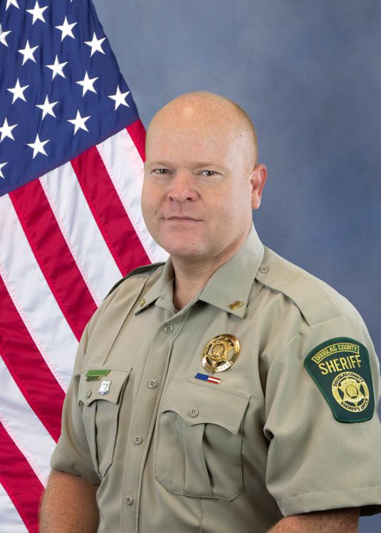 Portrait of a Douglas County sheriff officer in uniform standing in front of an American flag.