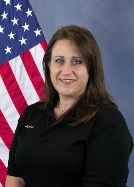 Portrait of a woman with shoulder-length brown hair wearing a black shirt with the name Jillian embroidered and standing in front of an American flag on a blue background.