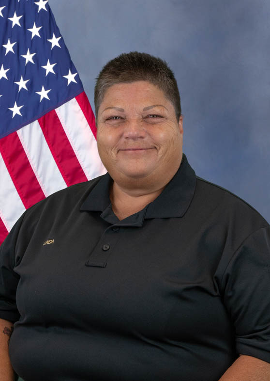 Portrait of a person wearing a black polo shirt with the name 'Linda' embroidered on it, posing in front of an American flag and a blue background.