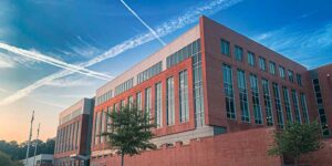 Exterior view of a modern multi-story brick and glass building under a blue sky with crisscrossing contrail lines at sunrise or sunset.