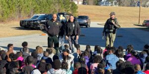 Three law enforcement officers standing and speaking to a large group of seated children outdoors with police vehicles in the background.