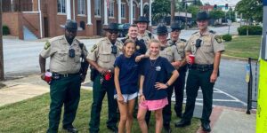 Group photo of seven uniformed sheriff officers standing on a sidewalk with two young girls in casual clothing, all smiling. The setting appears to be near a street intersection with brick buildings and greenery in the background.