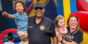 A smiling sheriff in a black shirt and cap holding a toddler, standing next to a woman and a young girl smiling in front of a colorful inflatable play structure.