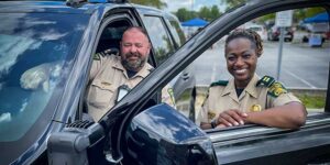 Two sheriff officers, a man and a woman, smiling while sitting in and leaning on a black vehicle in a parking lot.
