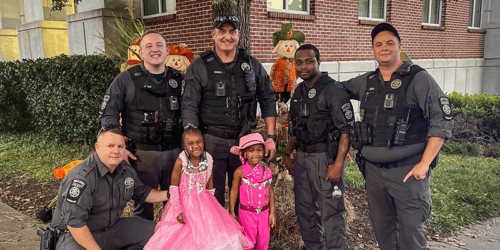 Group of six uniformed officers posing outdoors with two young girls dressed in pink Halloween costumes in front of Halloween decorations including scarecrows.