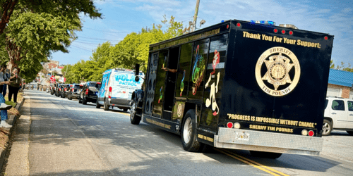 Sheriff's Department vehicle with music-themed decorations and the text "Thank You For Your Support: Sheriff Tim Pounds" and "Progress is impossible without change" in a parade procession on a tree-lined residential street with spectators.