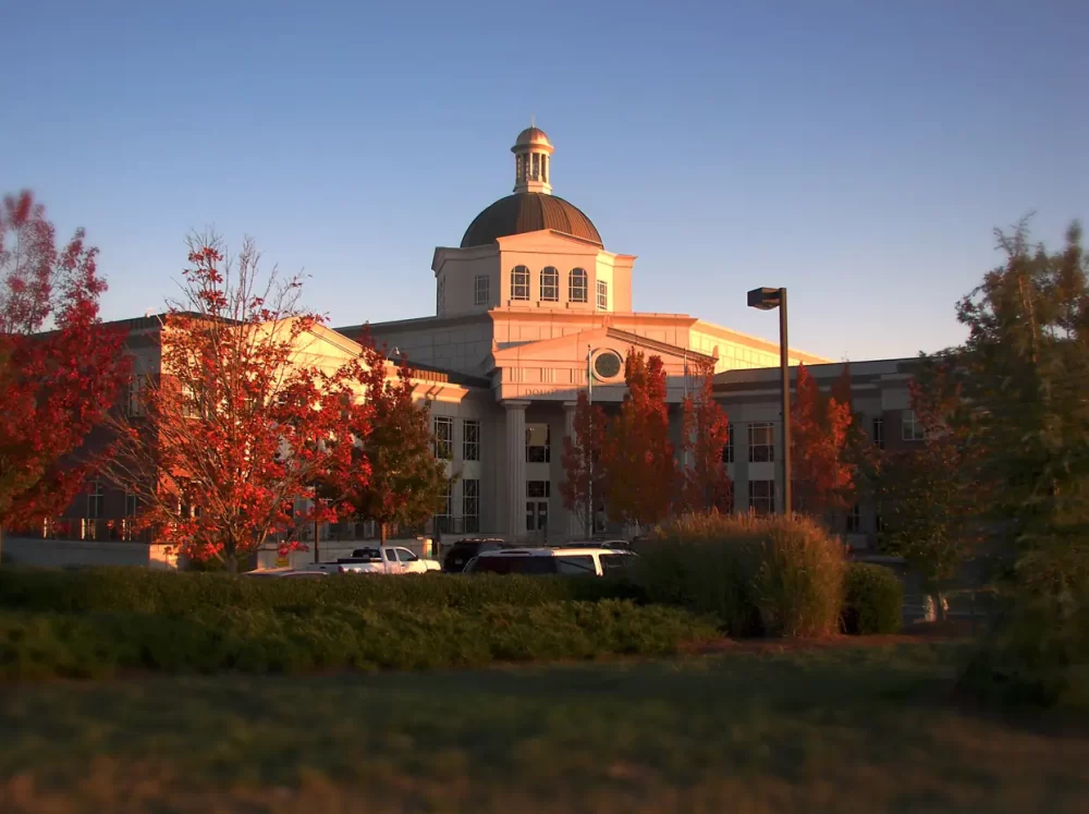 Courthouse building with a domed cupola and columns, surrounded by trees with autumn foliage and parked cars in front during sunset.